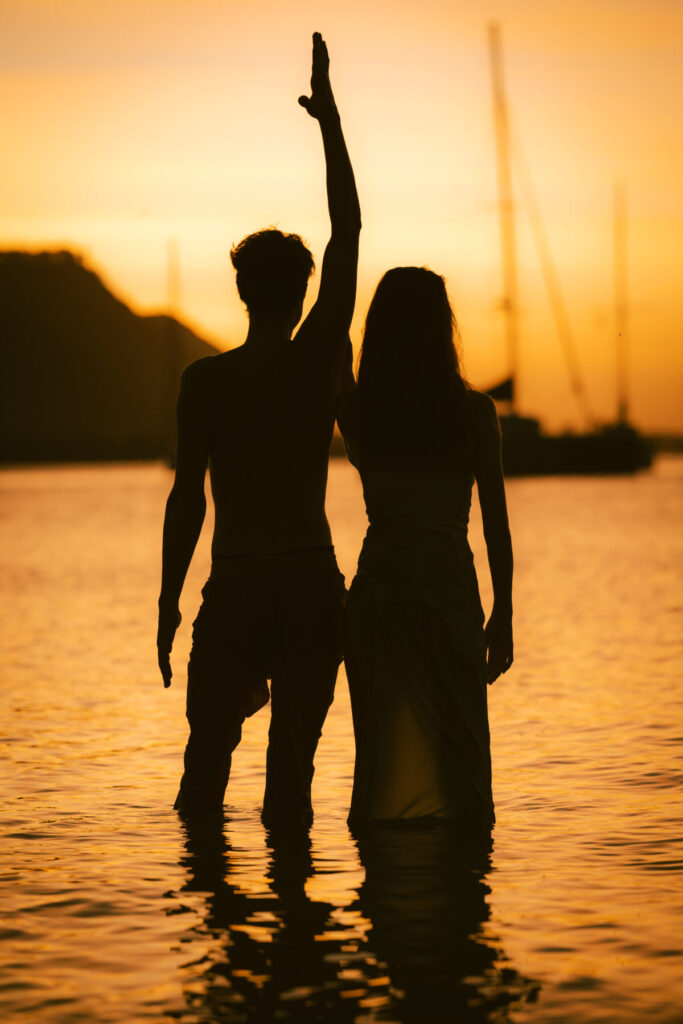 Spirituelle Befreiung bei Sonnenuntergang – Paar steht Hand in Hand im Wasser, Mann hebt den Arm, Licht voller Hoffnung und Freiheit