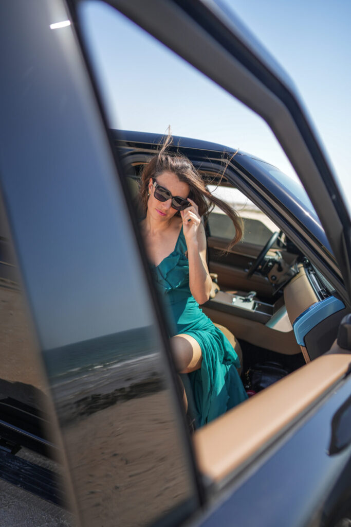 Beiträge mit Wirkung – Frau steigt elegant aus Auto am Meer, Sonnenbrille und Wind im Haar, Szene voller Stil, Ausdruck und starker Präsenz