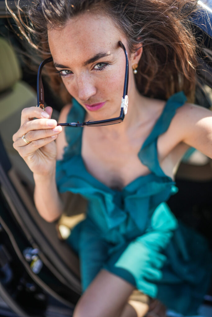 Leichtigkeit verkörpern – Frau im türkisfarbenen Kleid steigt aus dem Auto, hebt die Sonnenbrille, Blick in Bewegung, wach, spielerisch, klar