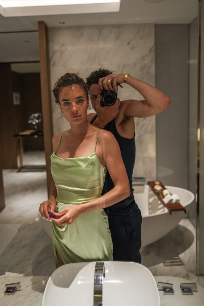 Erfolg teilen – Frau im eleganten Kleid, Mann mit Kamera, Spiegelmoment in stilvollem Bad, Symbol für Sichtbarkeit, Präsenz und Manifestation