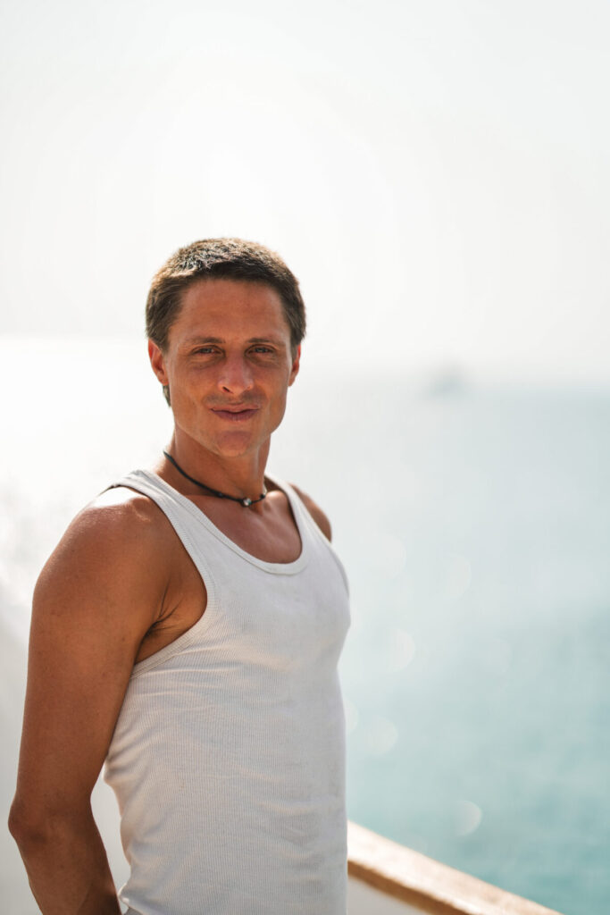 Spiegel der Liebe erleben: Mann im weißen Tanktop am Meer strahlt sanfte Verbundenheit und innige Twin-Flame-Intensität aus.