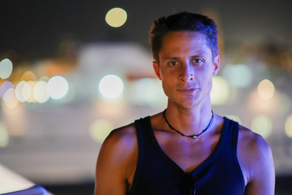 Langsame Reifung wählen – Mann im dunklen Tanktop vor unscharfem Stadtlichter-Bokeh, intensiver Blick und ruhige Präsenz strahlen geduldige Transformation aus.