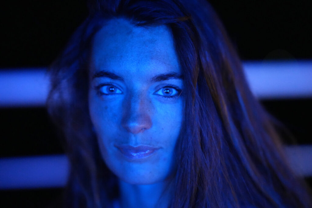 heilender Urton – Frau im blauen Lichtsturm schaut direkt und verkörpert die Gnade des kosmischen Ursprungsklangs in tiefem Gewahrsein.