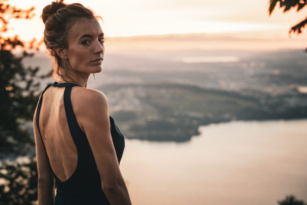 Mut zur Öffnung: Frau im schwarzen Kleid blickt über weite Wasserlandschaft bei Sonnenaufgang, kraftvoller Blick und Freiheit.