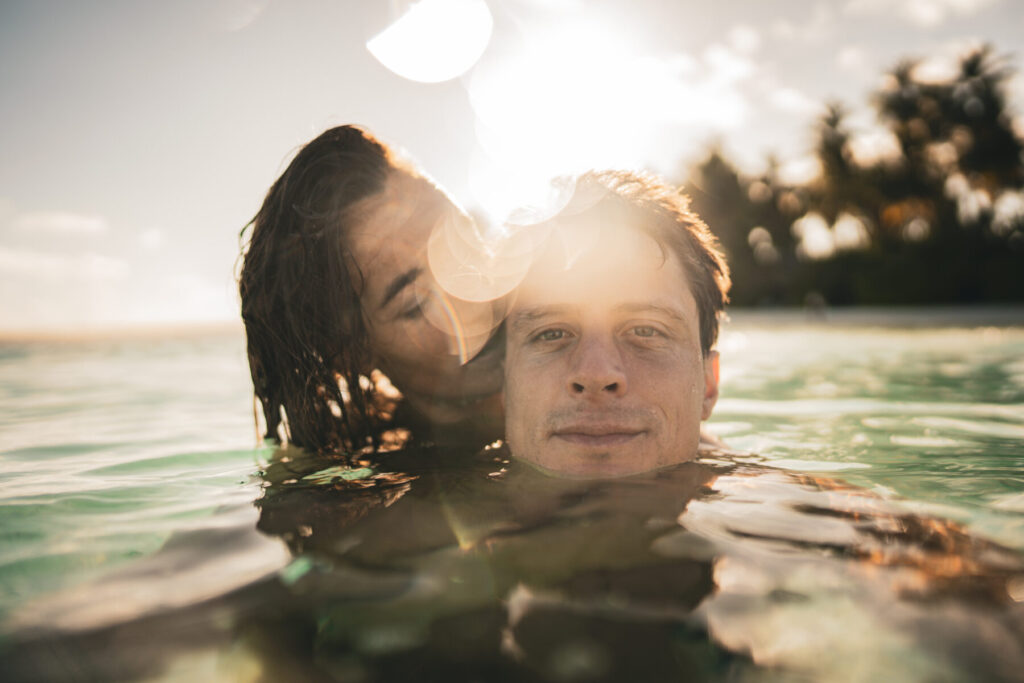 Verwundbarkeit offenbaren: Paar bis zur Brust im Wasser, Frau umarmt Mann im Nacken, Sonnenlicht strahlt als Wärme und tiefe Verbundenheit.