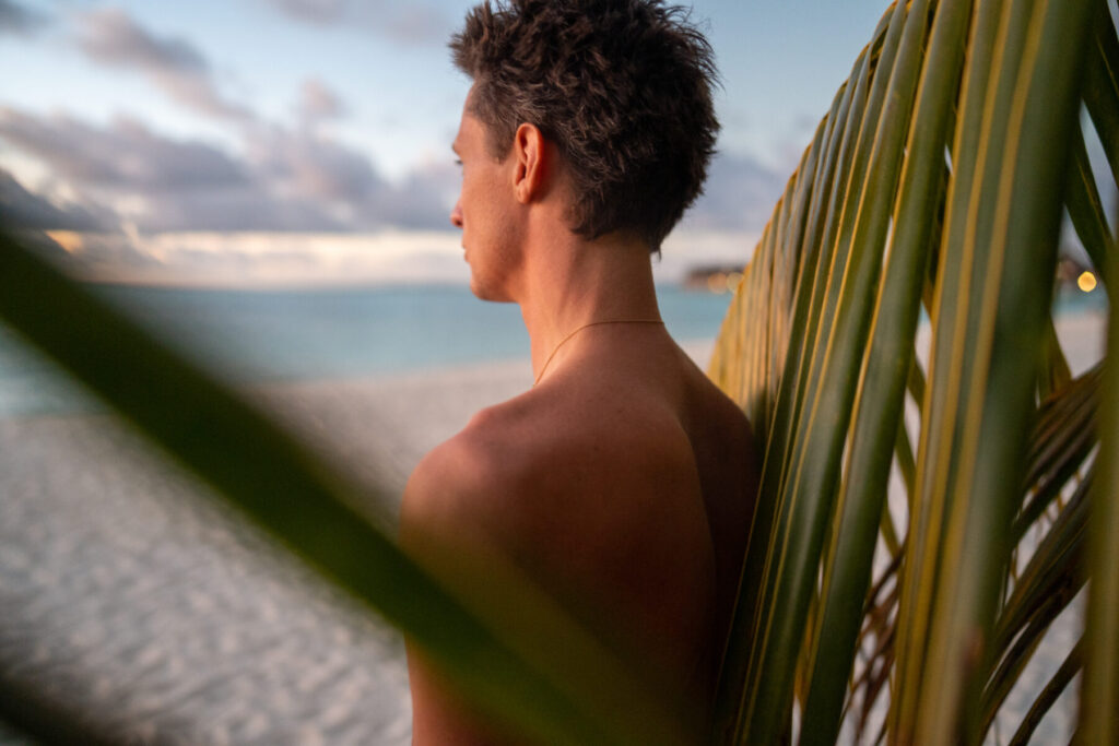 Bewusstseinsverbindung und Ruhe am Meer – Mann mit nacktem Oberkörper schaut über den Strand, teilweise verdeckt von Palmenblatt.