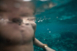 Reflektierende Fülle wählen – Halb untergetauchter Oberkörper im klaren Wasser, Wasseroberfläche teilt Perspektive und symbolisiert tiefes Eintauchen in deinen Wohlstand.