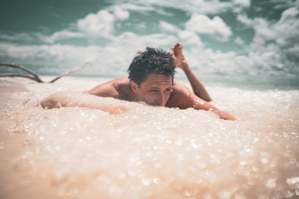 Authentische Offenheit wählen – Mann liegt auf Surfbrett im seichten Wasser, stützt sich auf Unterarme und blickt entschlossen voraus.