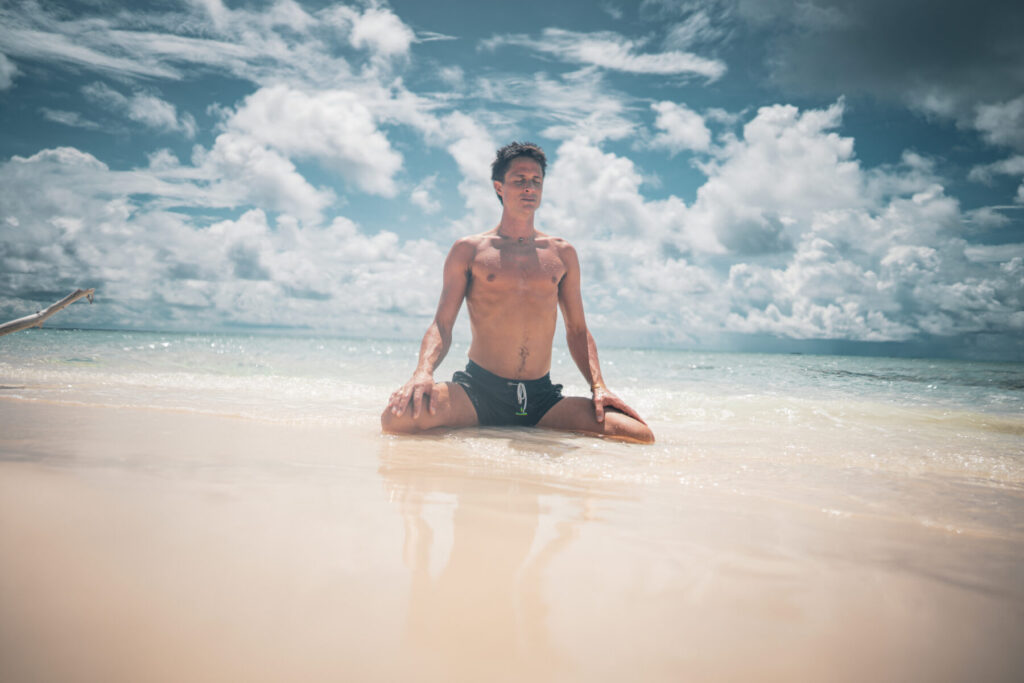 heilende Verschmelzung – Mann sitzt meditativ im flachen Meer, atmet tief ein und erlebt im Ozean-Tempel pure Liebes-Intimität.