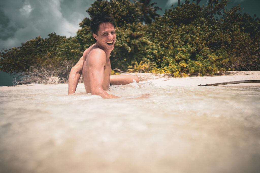 Herausforderung annehmen – Vater liegt im seichten Wasser am Strand, lächelnd in die Kamera blickend, spielerische Leichtigkeit und Stärke vereint.