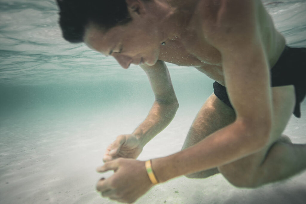 Vertrauen wählen – Mann taucht im klaren Wasser, Hände greifen sanft den Sandboden, strahlt ruhige Geborgenheit und harmonisches Miteinander.