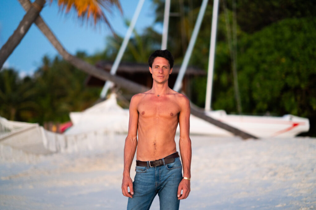 Wahre Freiheit wählen – Mann steht am Strand vor Katamaran, atmet ruhig und entlarvt Lügen, indem er in seine authentische Präsenz tritt.