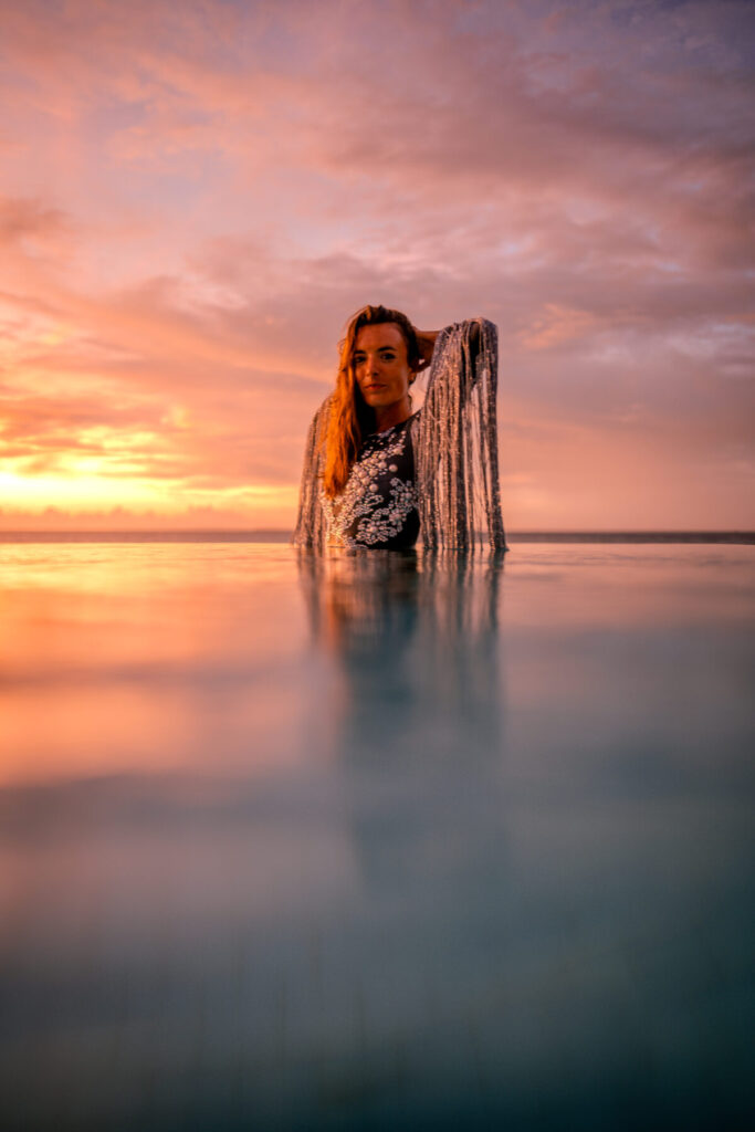 Magische Begegnung mit dem Licht – Frau steht im Wasser bei Sonnenuntergang, glitzerndes Kleid, Weite, Stille, goldene Präsenz und Sanftheit