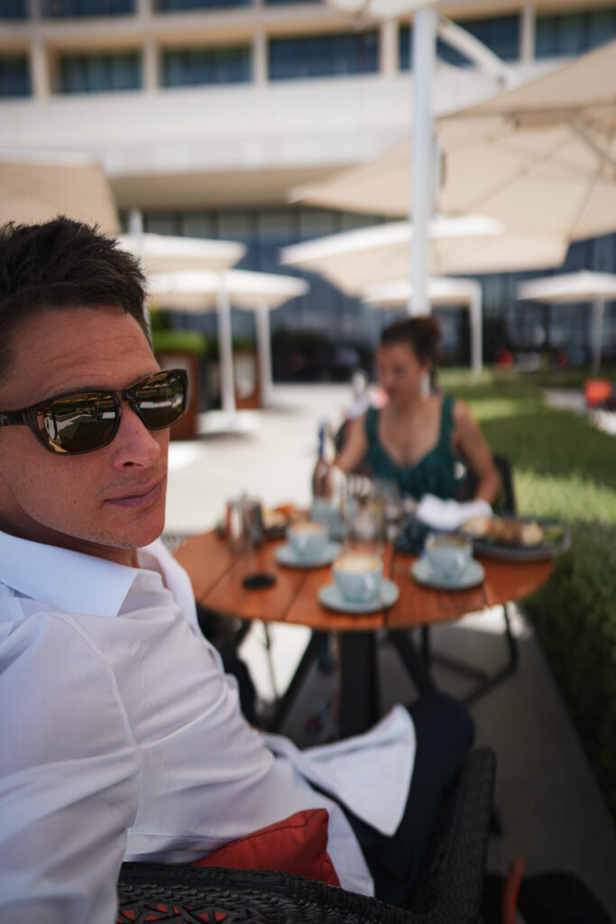 Den Moment ergreifen – Mann mit Sonnenbrille im Café, Frühstück im Freien mit Blick auf Partnerin, entspannte Fülle und gelebte Entscheidungskraft