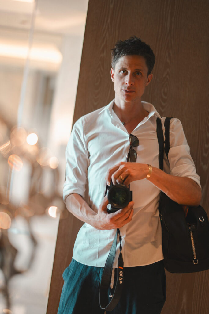 Business der neuen Zeit – Mann im weißen Hemd mit Kamera vor Hotellobby, Symbol für Präsenz, Kreativität und energetisch geführtes Unternehmertum