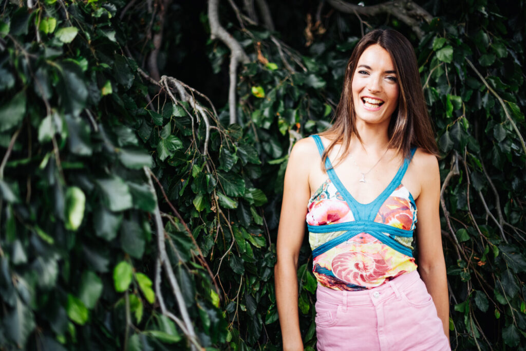 Frau steht lächelnd vor dichtem grünen Laub, trägt ein buntes Top mit Spiral-Muster und rosa Hose – Symbol für Gnade und die Gene Keys.