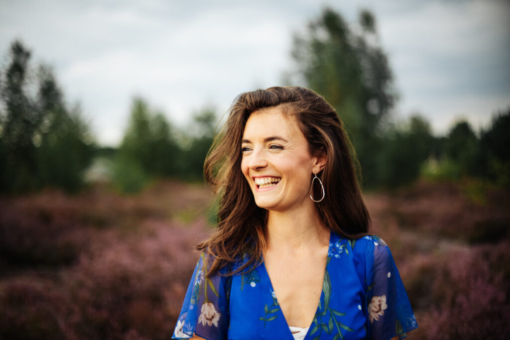 Geduldiges Wachstum wählen – Frau im blauen Kleid steht in lila blühender Landschaft, lächelnd den Blick zur Seite gerichtet, reife Präsenz spürbar.