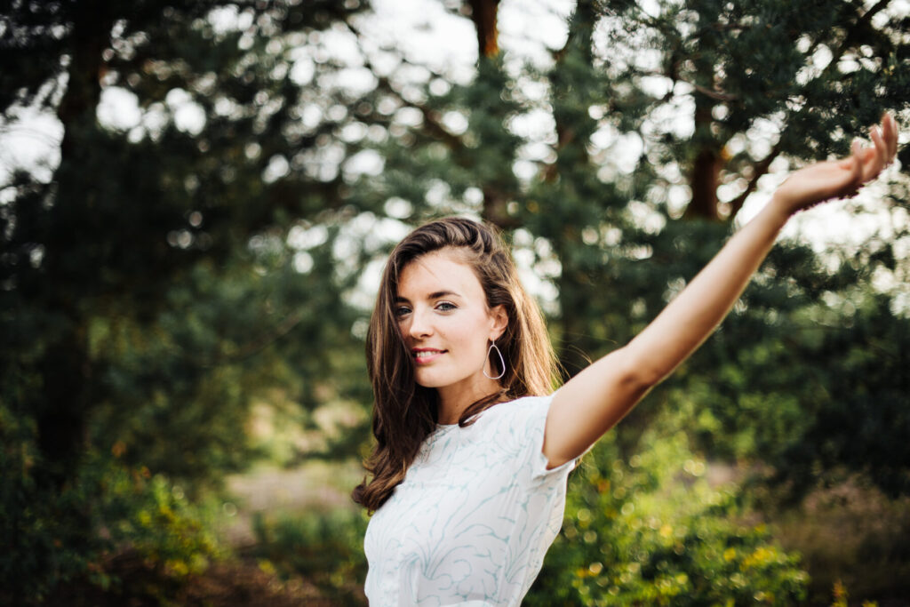 Entwicklung wählen – Frau in weißem Kleid mit erhobenen Armen vor grünem Hintergrund, lächelnd im Sonnenlicht, symbolisiert neuen Schritt und Wachstum.
