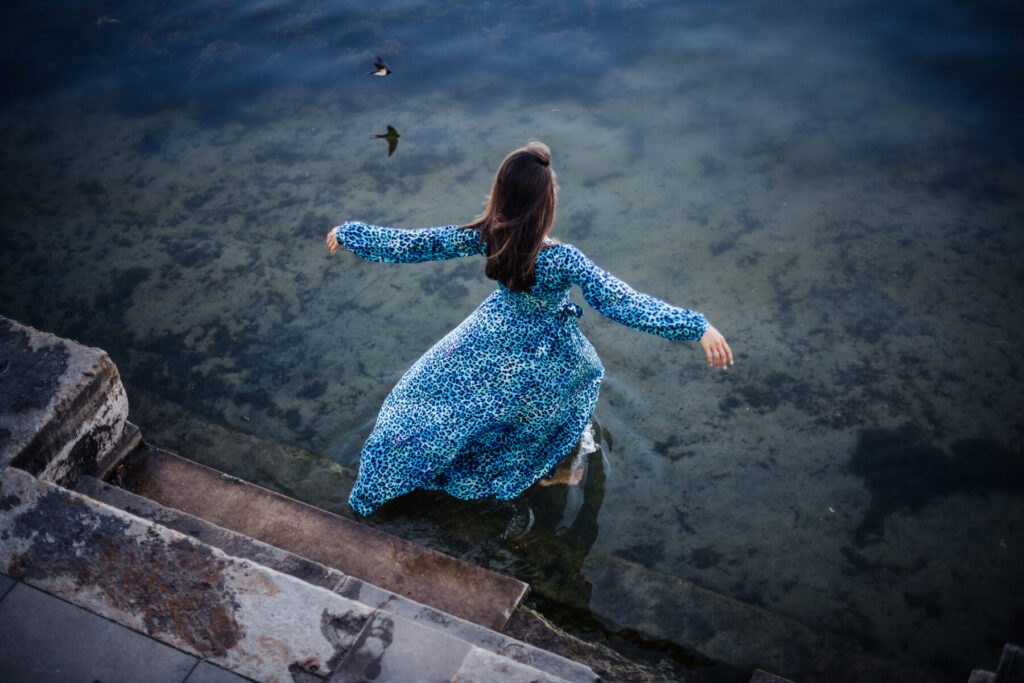 das Feld ruft – Frau im fließenden Kleid schreitet barfuß ins klare Wasser, offen, empfangend, verbunden mit Tiefe und heiligem Moment