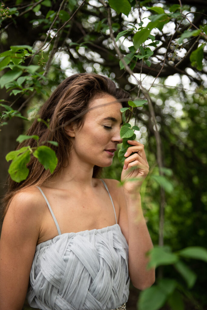 Ressourcenbewusstsein wählen – Frau im grauen Kleid hält ein Blatt vor der Nase, genießt achtsam die Natur und symbolisiert nachhaltige Fülle.