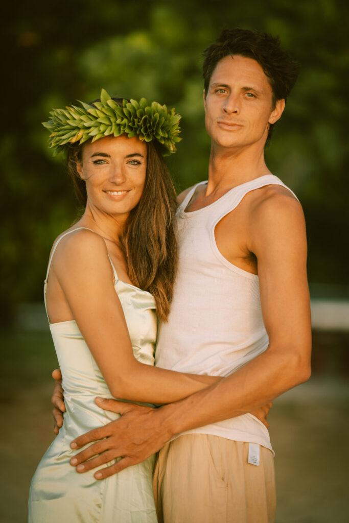 Heilung empfangen – Paar in weiß am Strand, Frau mit Blumenkranz, strahlt Anbindung, Erdung und liebevolle Präsenz im Lichtfeld aus