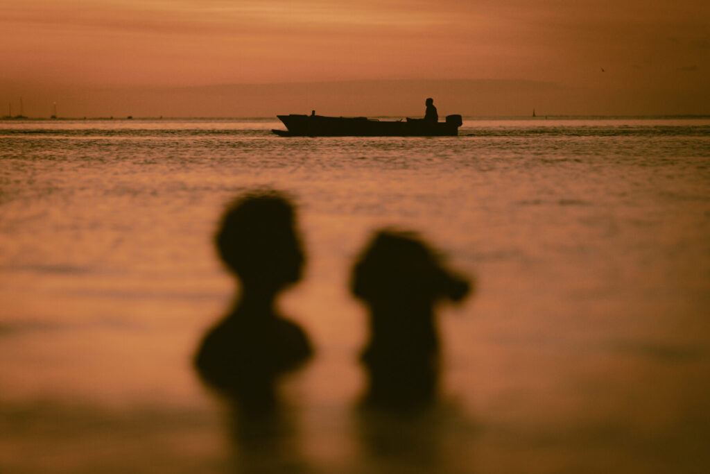 Langsame Entfaltung wählen – Unscharfe Silhouetten zweier Personen im seichten Wasser vor einem Boot bei Sonnenuntergang, Symbol für geduldiges Wachstum und Beständigkeit.
