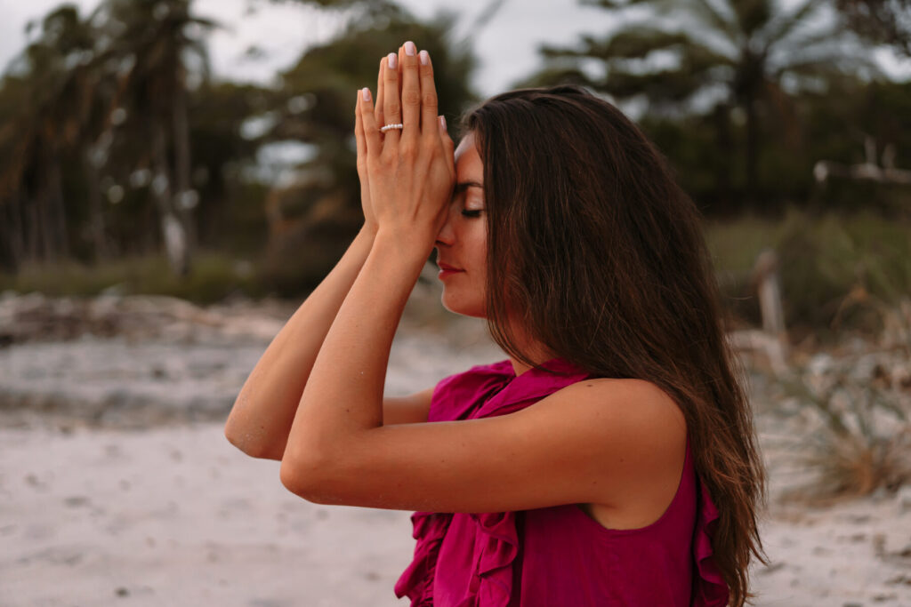 Innere Ruhe wählen – Frau im pinken Kleid steht am Strand, Hände vor dem Gesicht gefaltet und Augen geschlossen, strahlt meditative Gelassenheit im Angesicht der Angst.