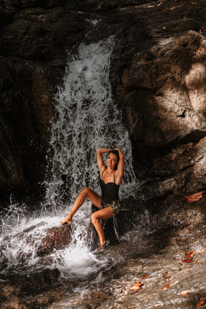 Harmonie im Moment wählen – Frau sitzt nackt unter einem Wasserfall, erhobene Arme und geschlossene Augen strahlen vollendete Verbundenheit mit sich selbst aus.