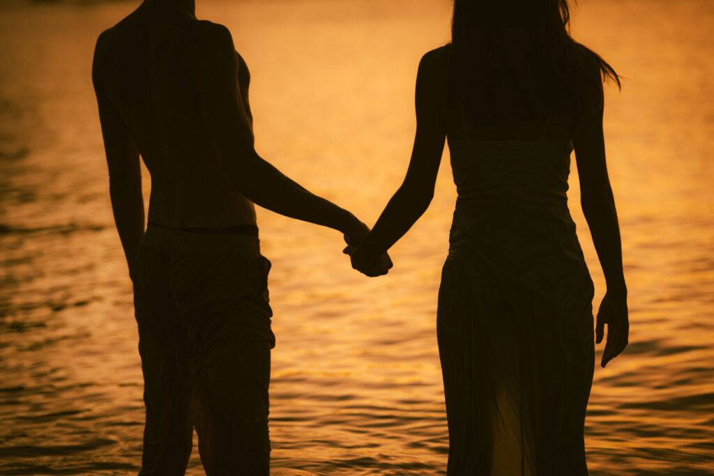 Zwei Menschen halten sich an den Händen im goldenen Licht des Meeres – Symbol für Verbindung, Vertrauen und das Thema Liebe macht sehend.